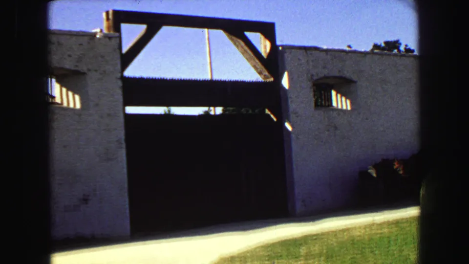 A visitor-shot 1968 8mm view of a white stucco wall and large dark gate with a small arched window, lawn, and bright sidewalk.