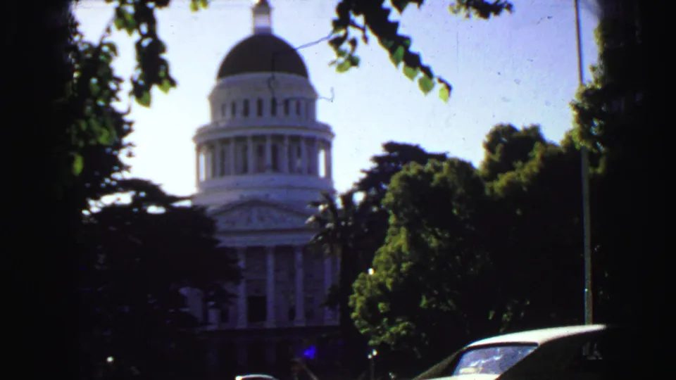 1968 8mm frame showing a domed white civic building partly hidden by leafy trees, with a flagpole and parked car at street level.