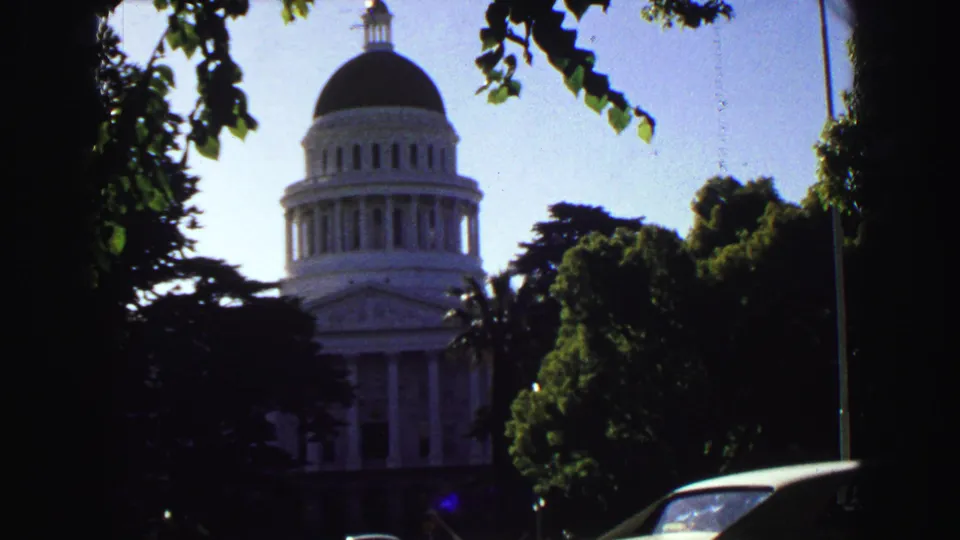 A late clip frame shows the domed white building framed by trees, a flagpole, blue sky, and the roofline of a parked car.