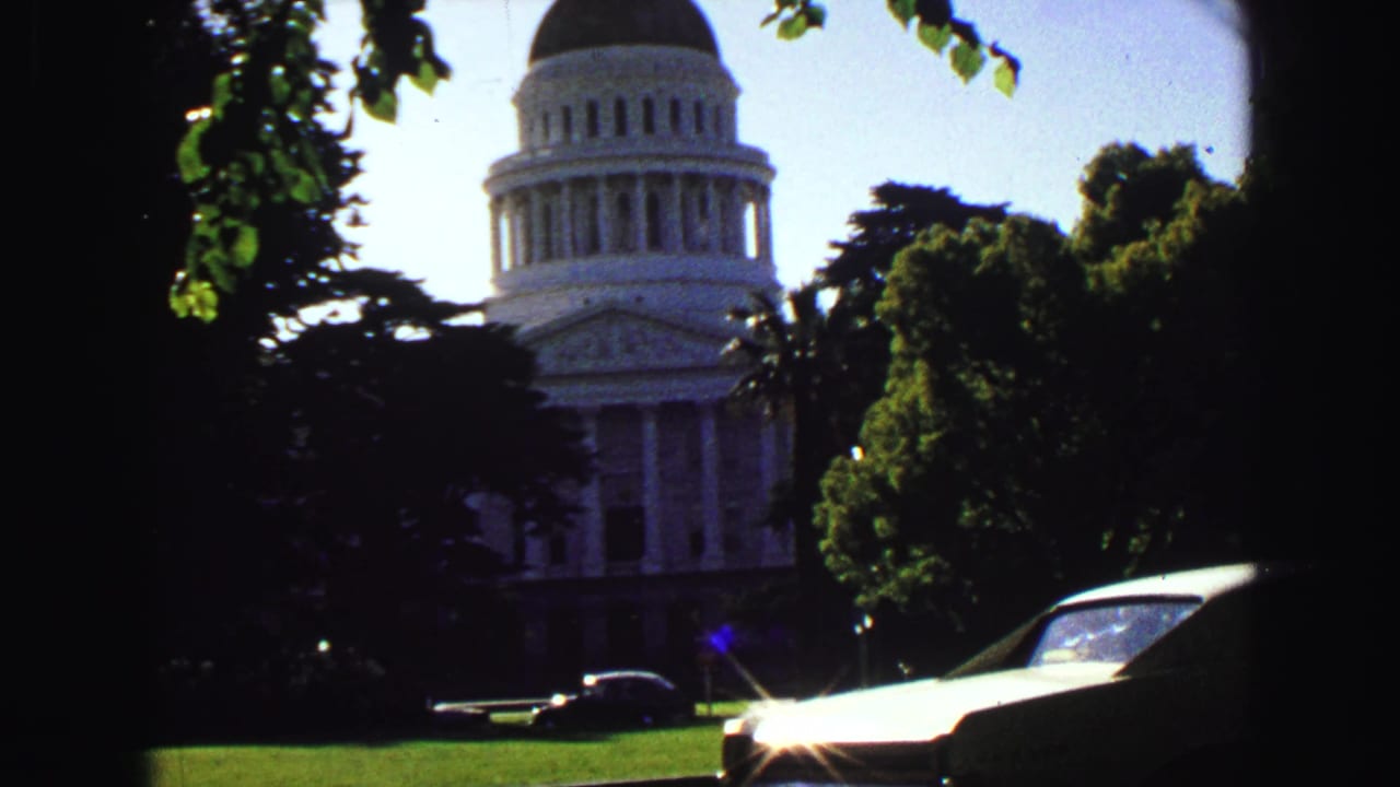 White domed civic building behind shaded trees, with lawn and mid-century cars visible in the foreground.