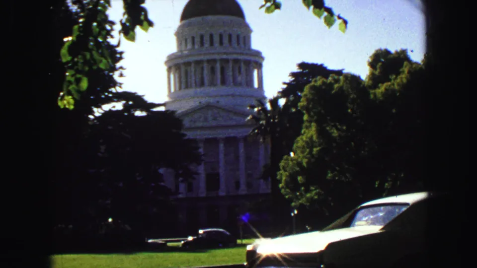 White domed civic building behind shaded trees, with lawn and mid-century cars visible in the foreground.