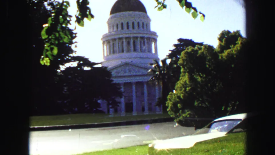 1968 home-movie frame showing a white domed civic building through trees, with lawn, street edge, and classic cars.