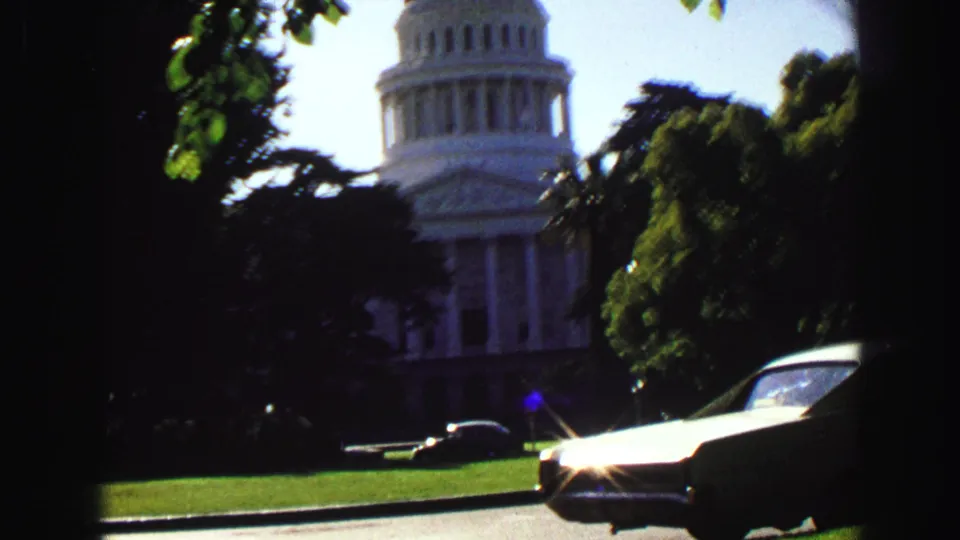 Domed civic building partly obscured by trees, with classic cars, lawn, and a bright flare on chrome in front.