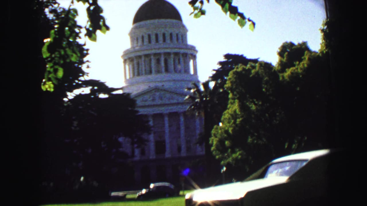 White domed civic-style building partly framed by trees and a passing car.