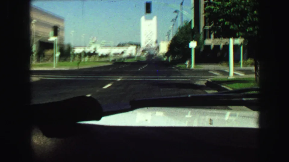 1968 home-movie windshield view along a city street toward a distant tower-like bridge structure and roadside buildings.