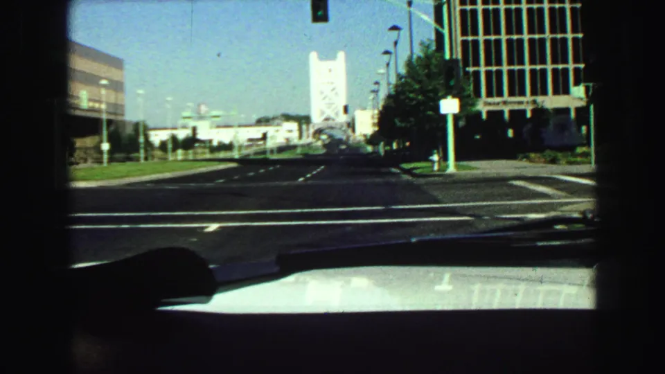 1968 in-car street view with lane markings, traffic lights, modern roadside buildings, and a distant tower-like bridge form.