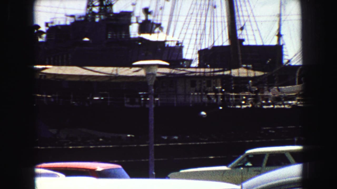 Moored vessels with tall masts behind parked cars at a waterfront.
