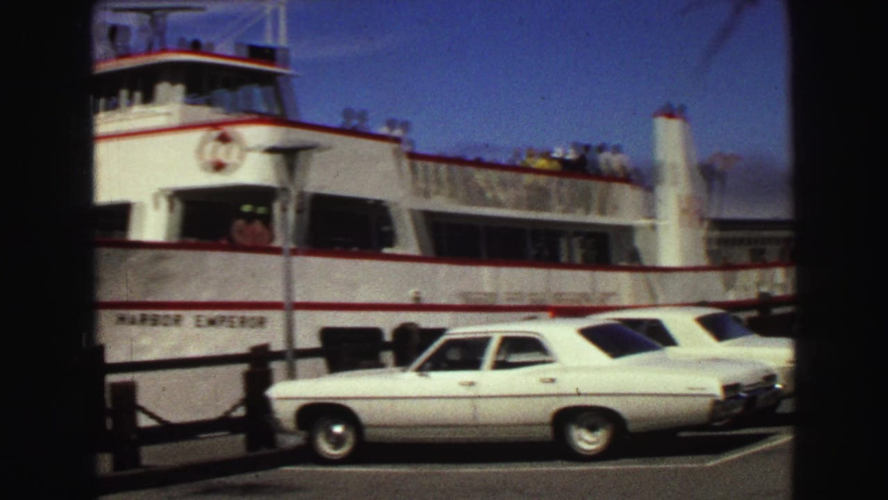 1968 Vancouver home-movie frame showing a white passenger ferry docked beside classic sedans near a pier.