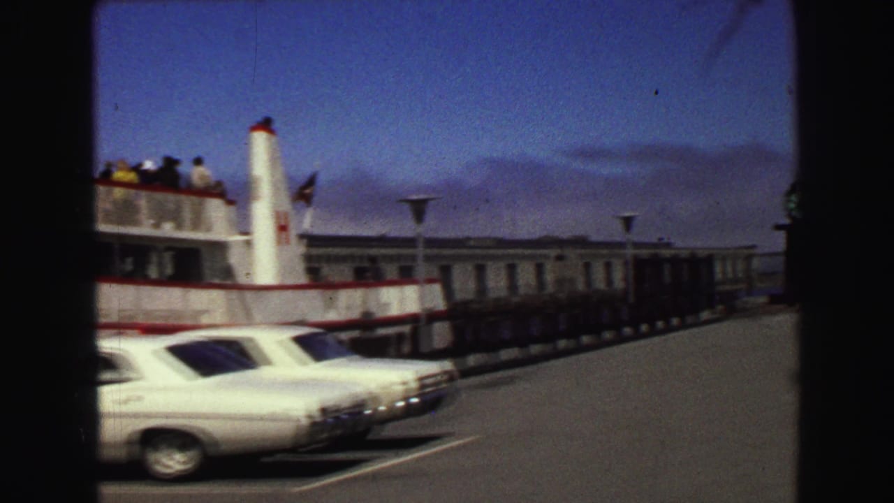 Passenger ferry, parked cars, and long pier building under blue sky.