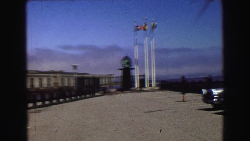 1968 Vancouver waterfront plaza with flagpoles, a clock monument, parked sedans, and a lone pedestrian in the distance.