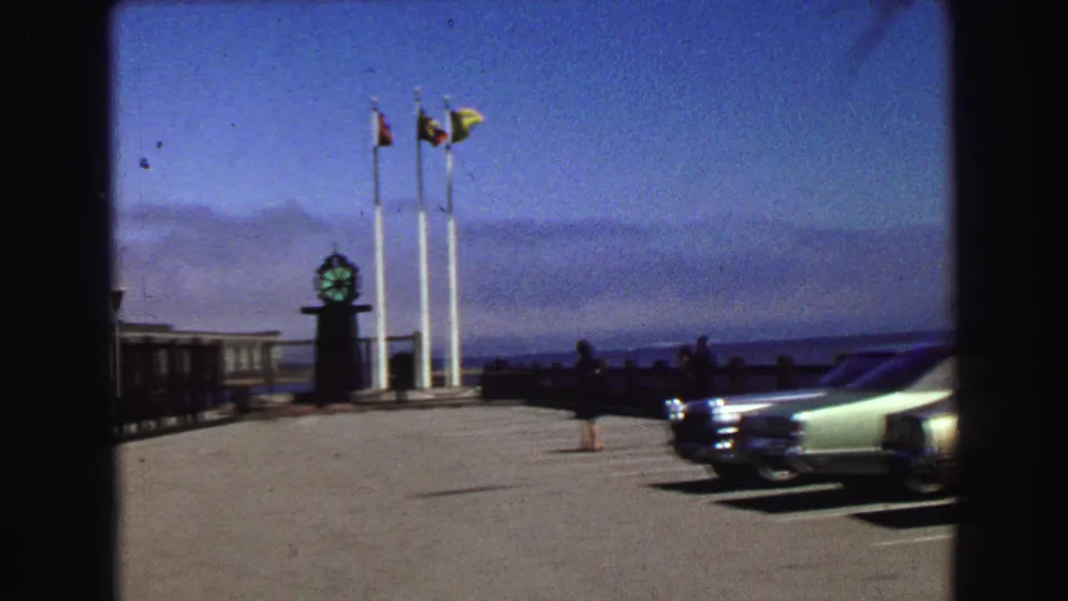 1968 Vancouver waterfront frame with flags, a clock monument, parked sedans, and distant people near the pier edge.