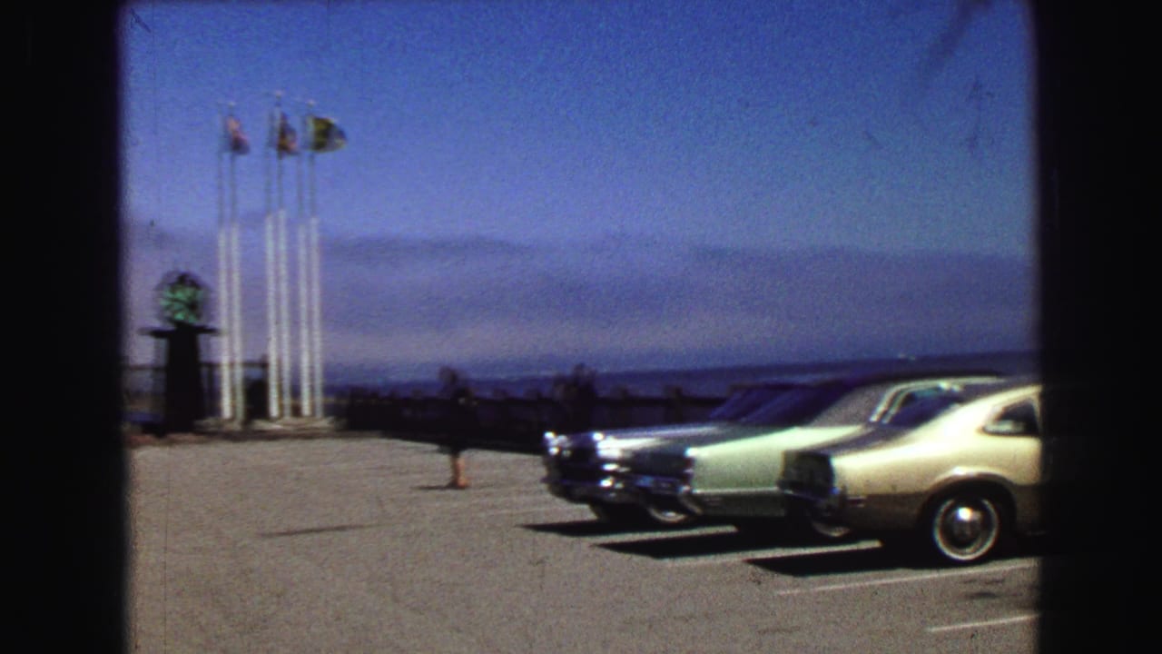 Parked cars face a waterfront parking lot with flagpoles and pedestrians at left.