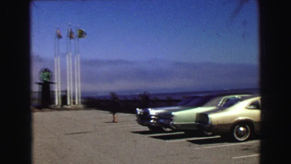 1968 home-movie frame of classic cars in a waterfront lot, with flagpoles, pedestrians, and open water beyond.