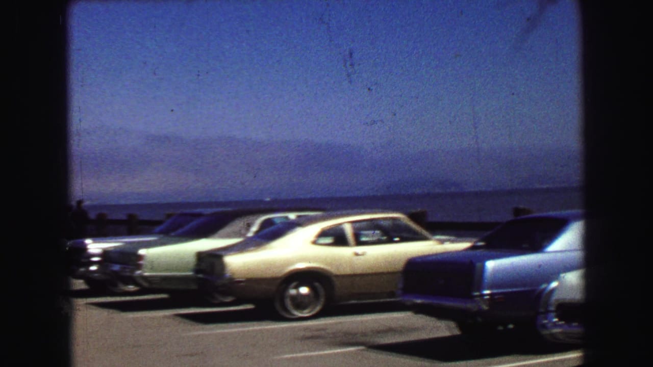 Cream coupe and other cars parked beside open water under a blue sky.