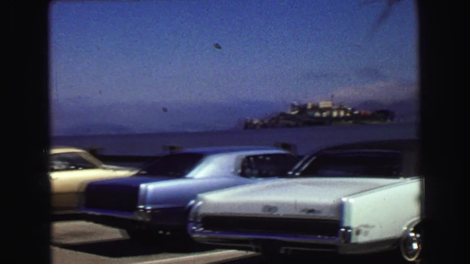 1968 home-movie frame showing parked cars at a waterfront lot and a distant island across the bay.