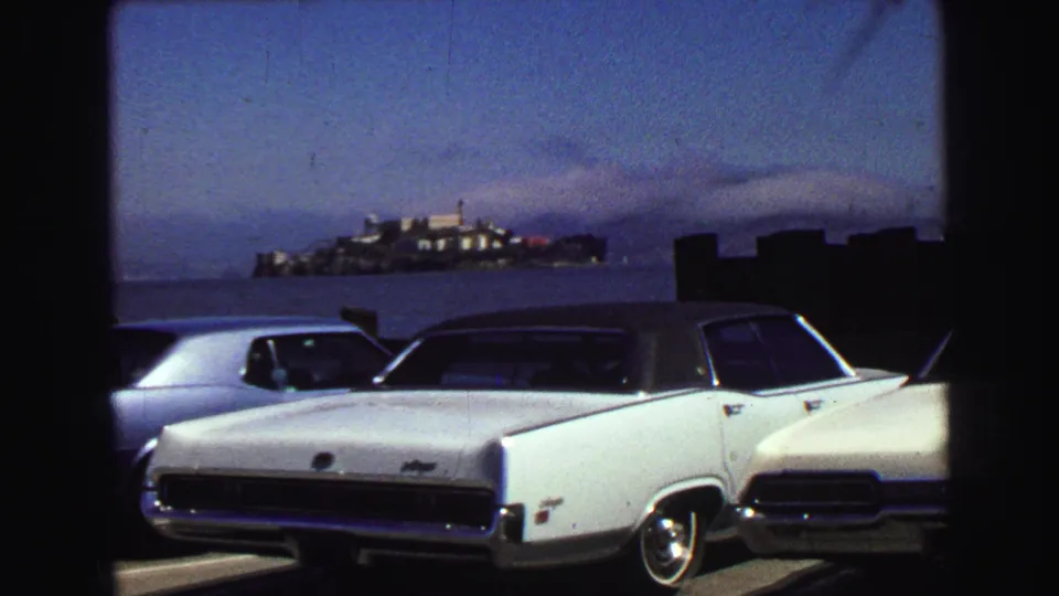 1968 waterfront parking scene with a white sedan in front and a distant island landmark across the water.