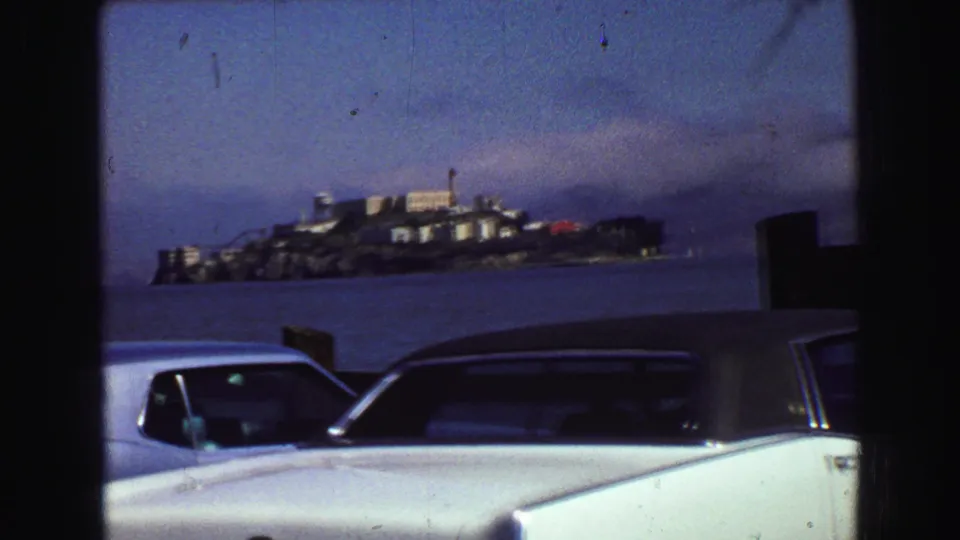 1968 8mm view toward Alcatraz Island across San Francisco Bay, with late-1960s cars parked along the waterfront.