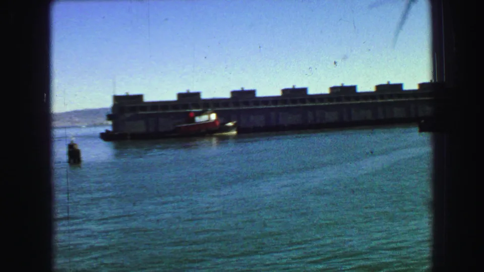 A long pier or waterfront building stretches across San Francisco Bay, with a small boat and distant shoreline visible in the 8mm frame.