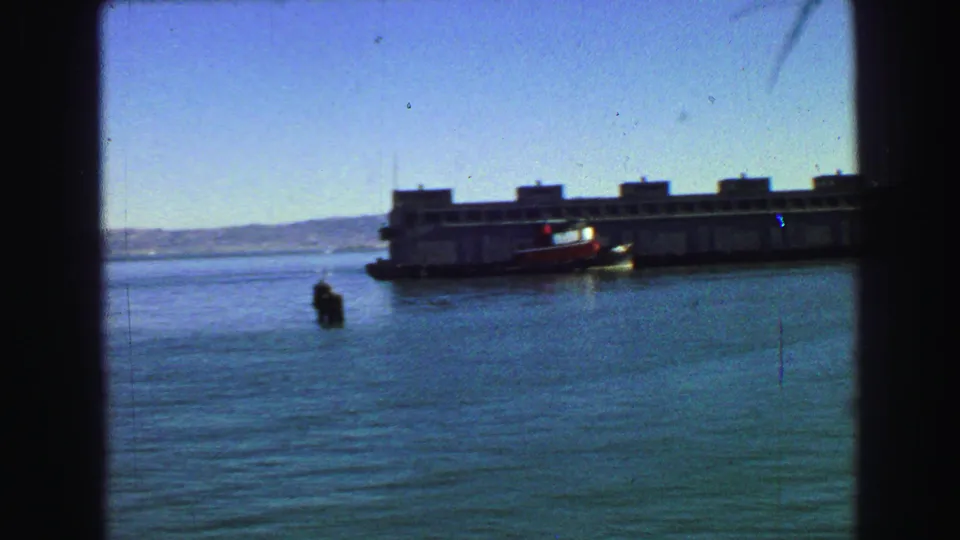 1968 8mm waterfront view from a moving vessel, showing a large barge or ferry-like craft, small boat, calm water, and low hills.