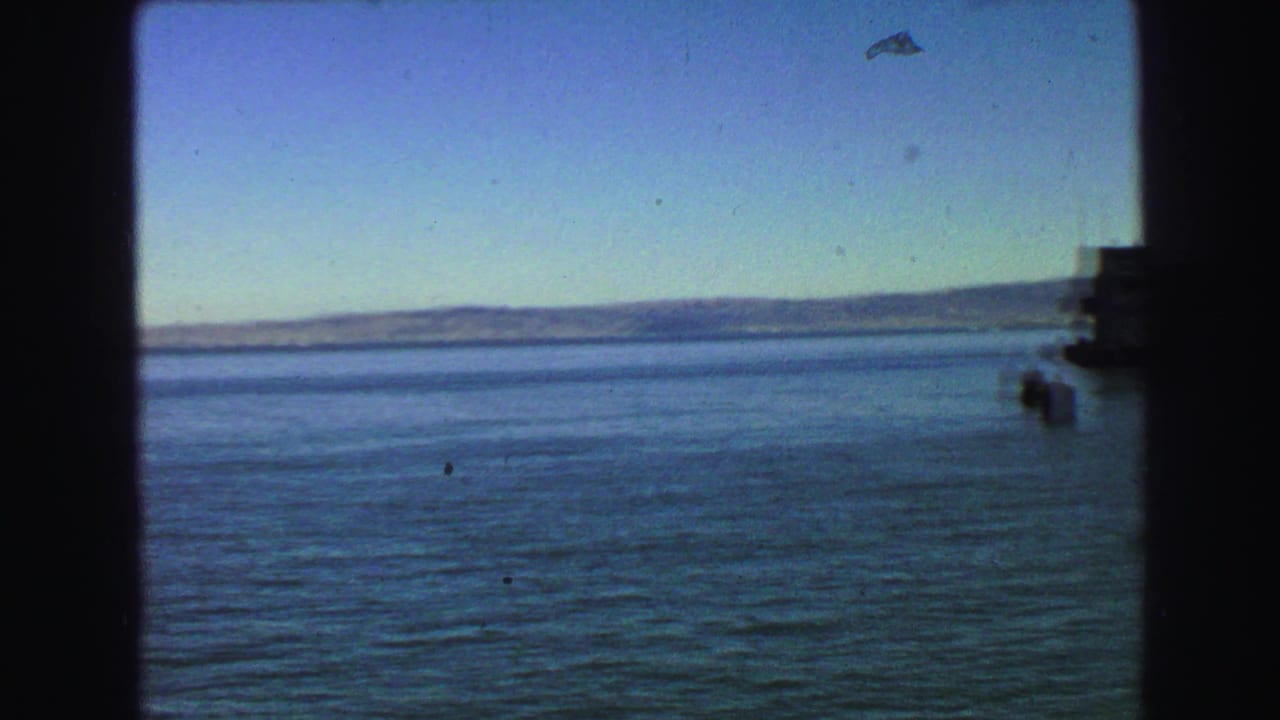 1968 8mm moving-vessel view across open blue water toward a low distant shoreline, framed by dark interior window edges.