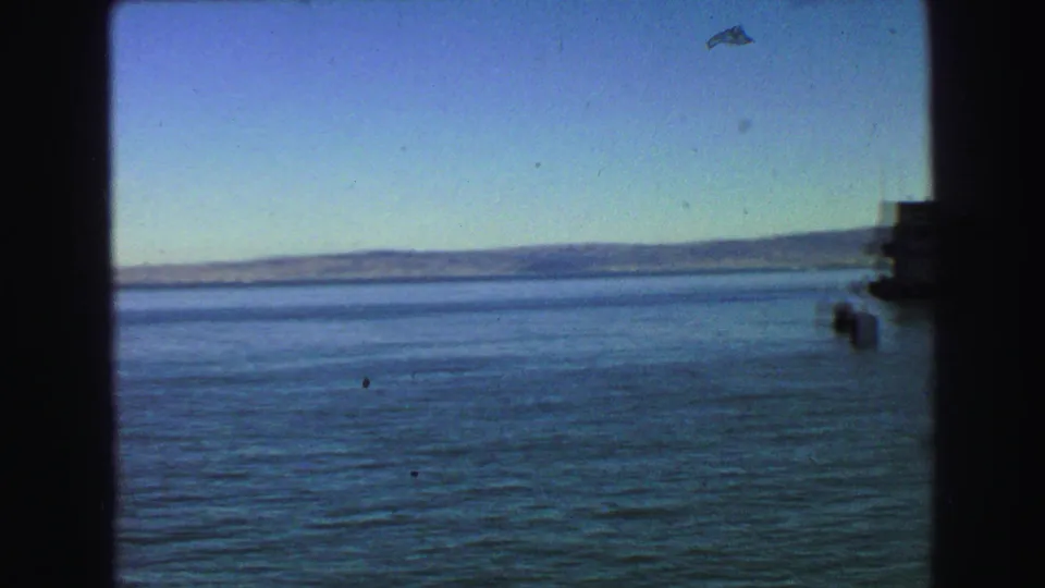 1968 8mm moving-vessel view across open blue water toward a low distant shoreline, framed by dark interior window edges.