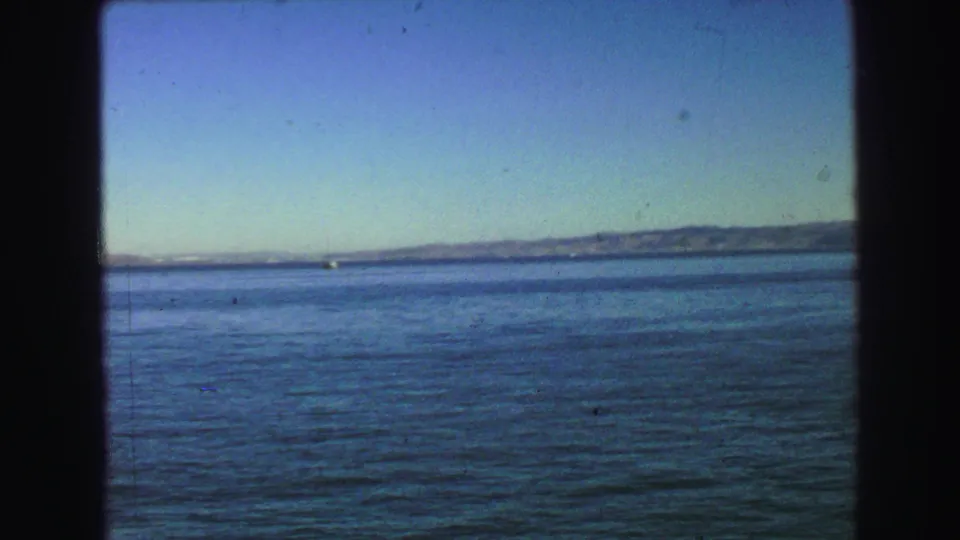 1968 8mm harbor panorama showing calm water, a tiny white boat near the horizon, and distant hills under a clear sky.