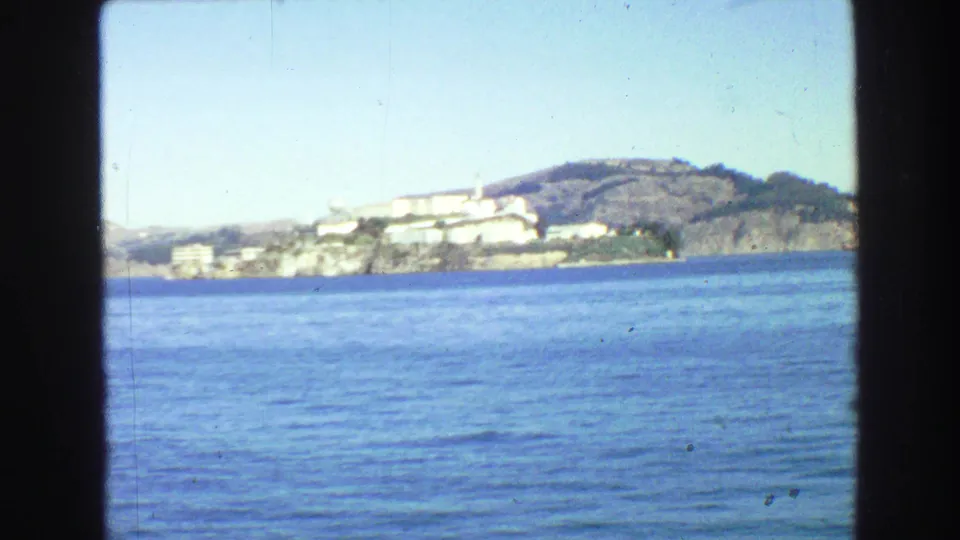 1968 8mm waterfront view of a rocky island with white buildings across blue water, consistent with the clip's Alcatraz identification.