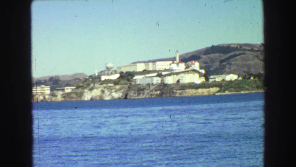 A slightly tilted 8mm frame shows Alcatraz Island across San Francisco Bay, with pale buildings above a rocky shoreline.