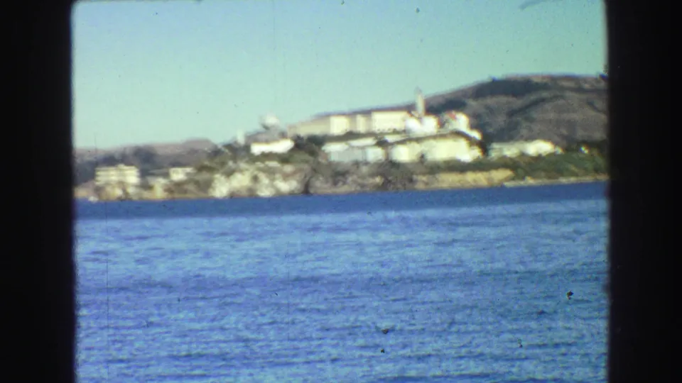Soft-focus 1968 8mm view of Alcatraz across blue bay water, showing the white complex, shoreline, and hillside backdrop.
