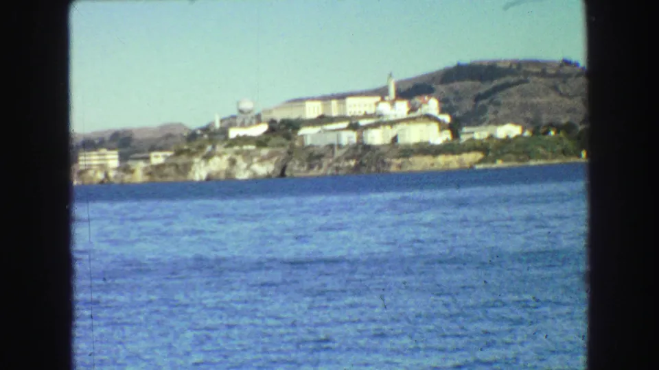 1968 8mm travel view across San Francisco Bay toward an island shoreline with buildings, framed by dark film edges.