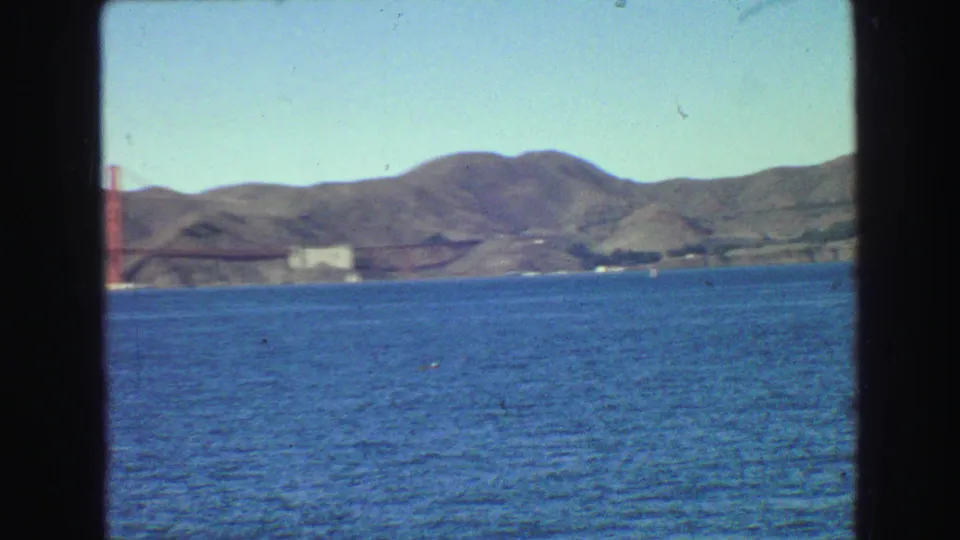 Distant 1968 home-movie view of the Golden Gate Bridge over bay water, framed by dark vertical film borders.