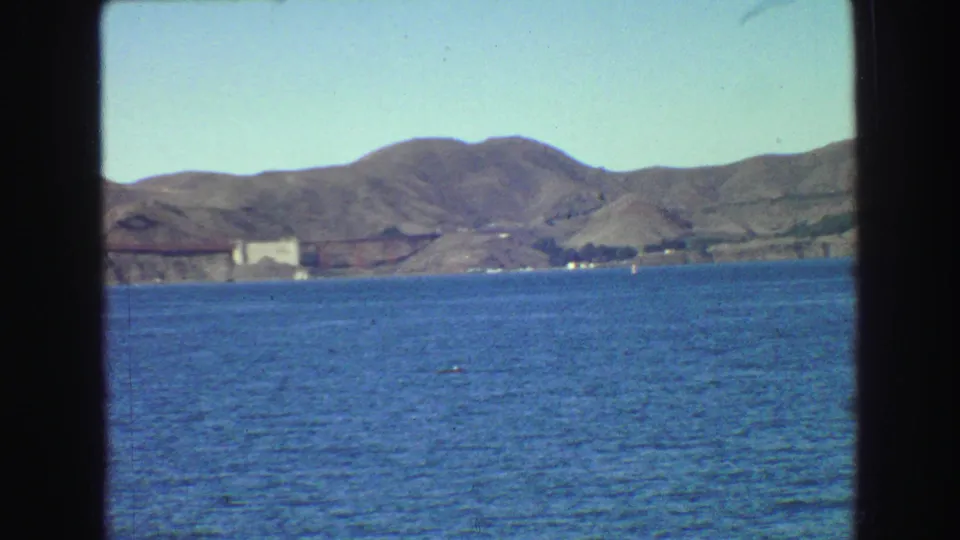 Soft 8mm frame of the Golden Gate Bridge area from the water, with bridge structure, hillside, and shoreline in the distance.