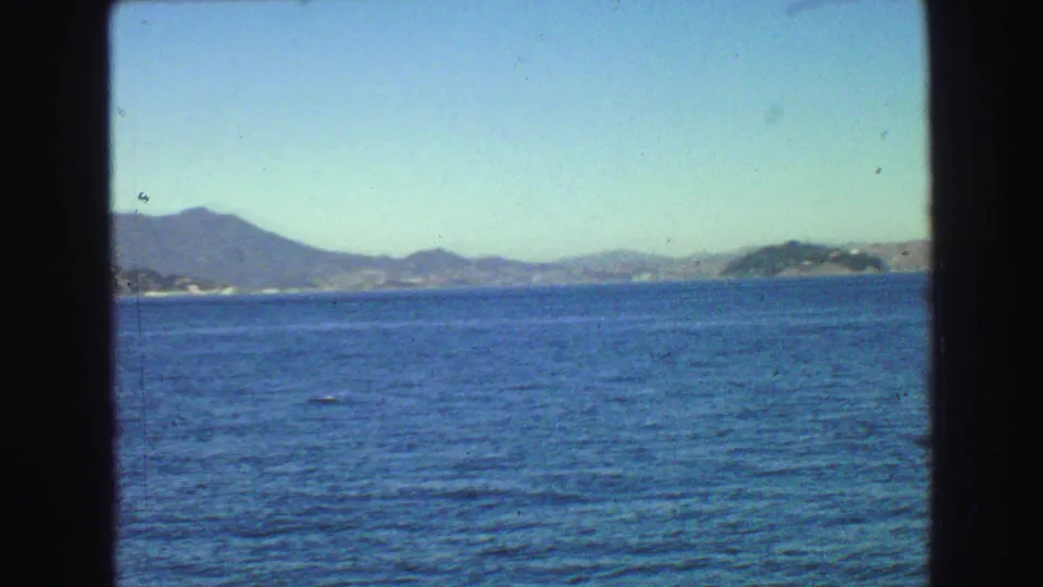 1968 Vancouver-area 8mm view across rippled water to distant hills and shoreline, with boat-window framing visible.
