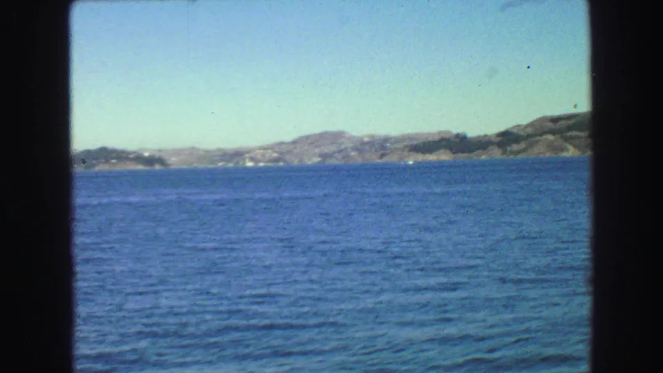 1968 Vancouver-area 8mm boat-window view of blue water, distant hills, and a developed shoreline under a clear sky.