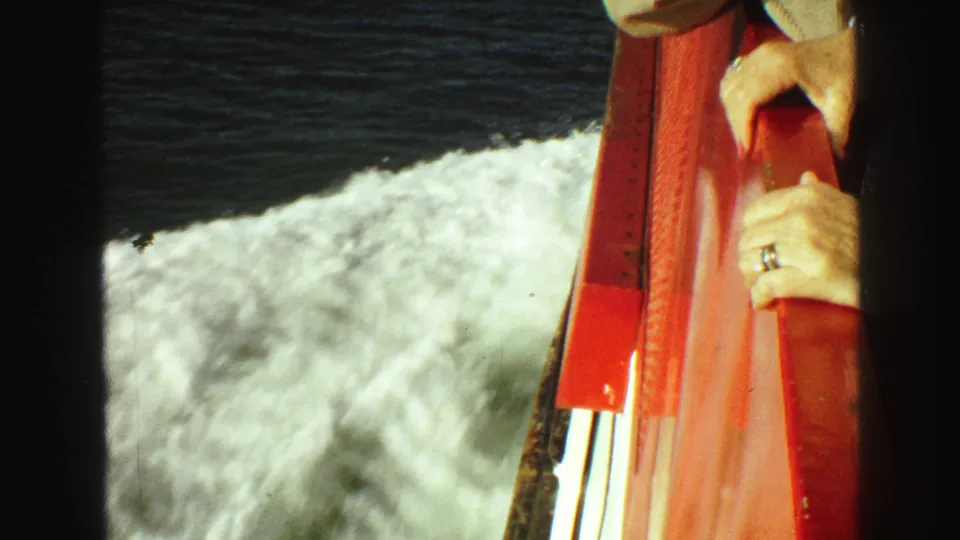 1968 8mm boat-view frame near Vancouver showing foamy wake, red railing, and a passenger's hands at the side.