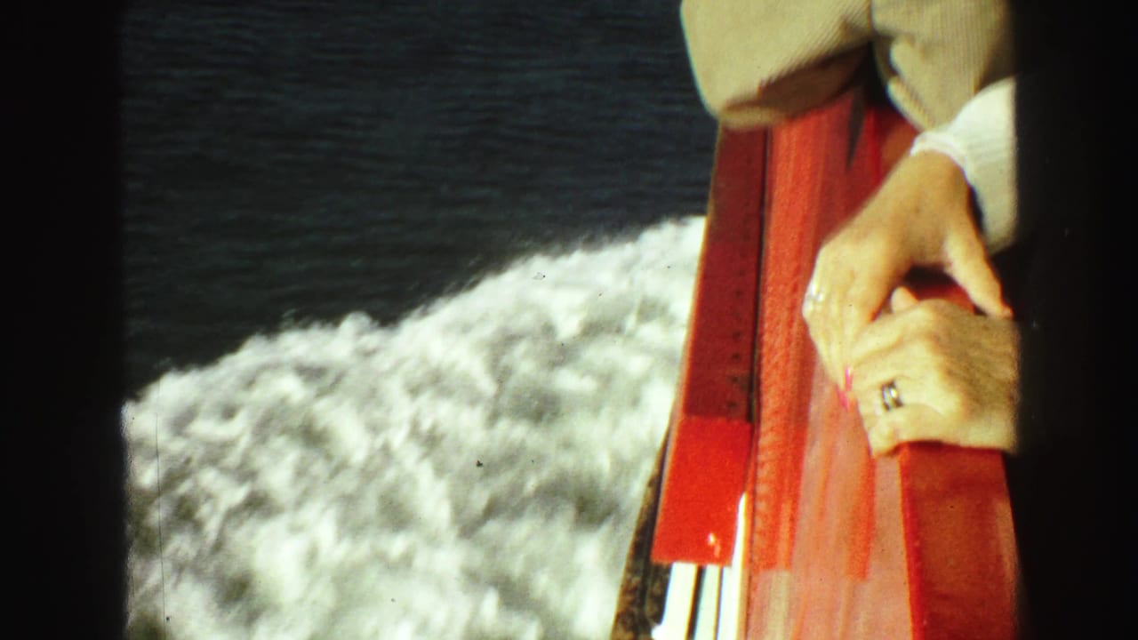 Close view of ringed hands holding a red ship railing above foamy water.
