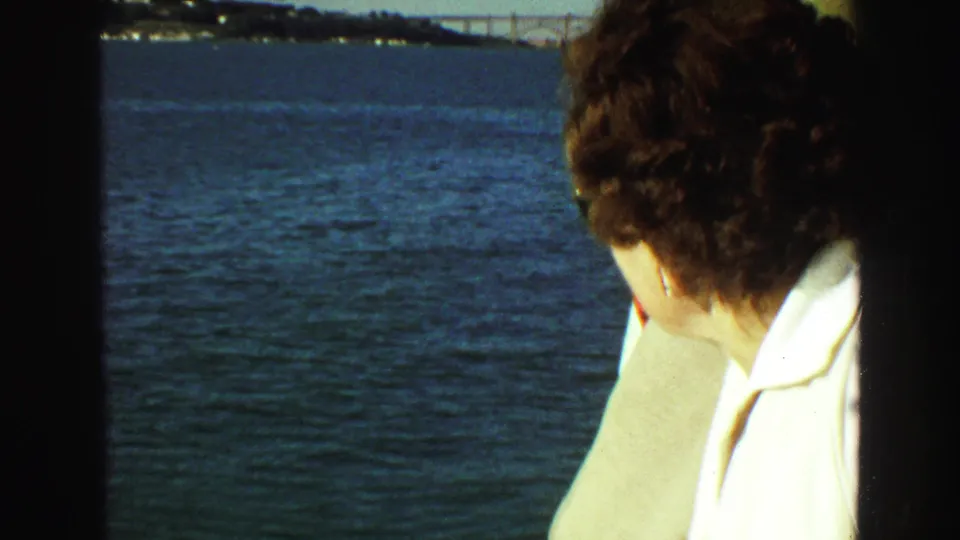 From behind, a woman in a white shirt and beige jacket looks across the water toward a distant Vancouver shoreline.