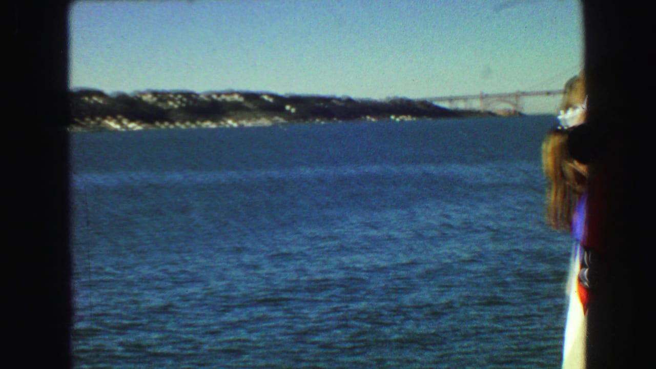 Ferry view over blue water with distant hills, bridge and a woman partly visible at the right edge.