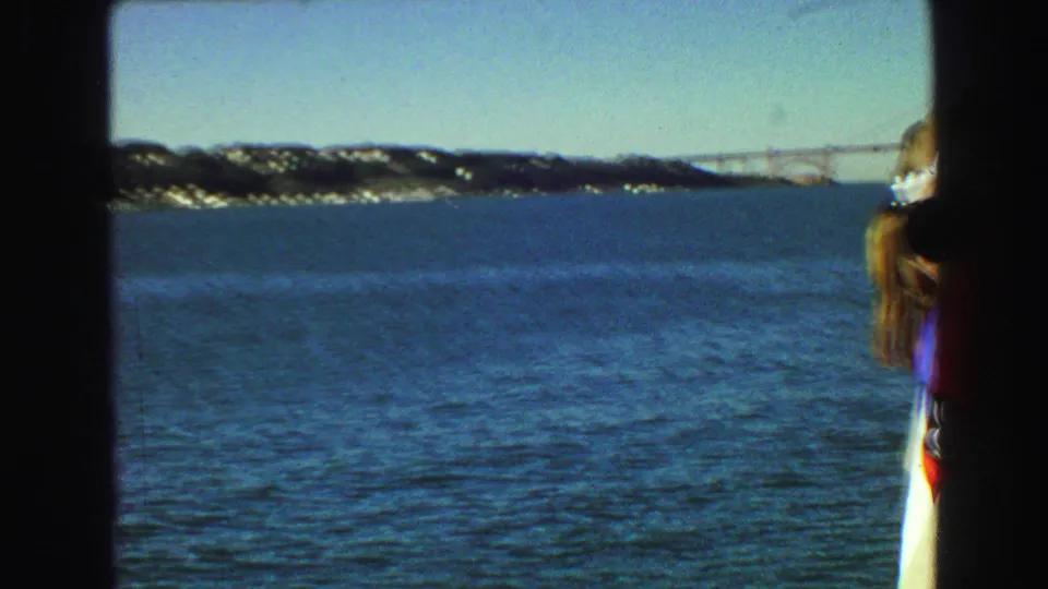 1968 Vancouver ferry view over blue water toward a distant bridge and shoreline, with a passenger partly visible at the frame edge.