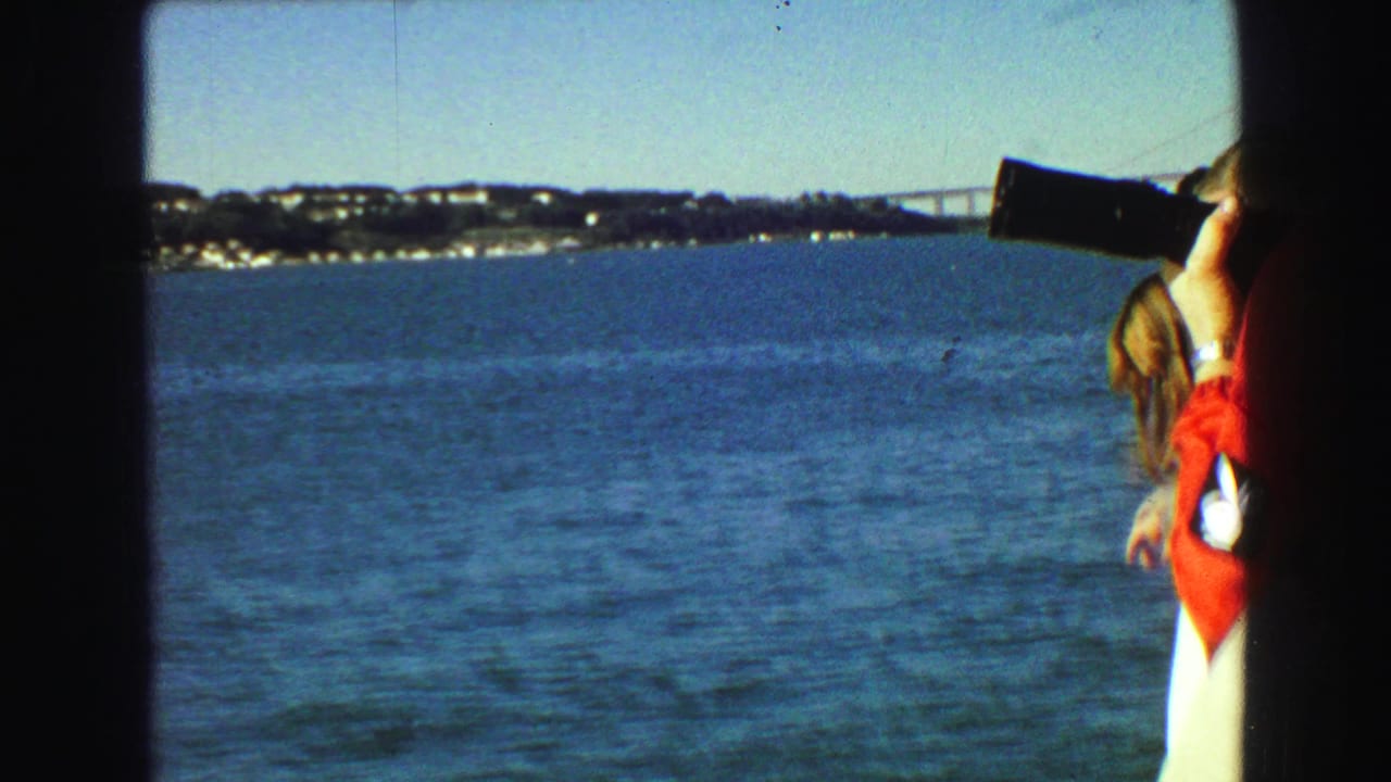 Woman in a red jacket and white hat raises a camera while looking across open water.