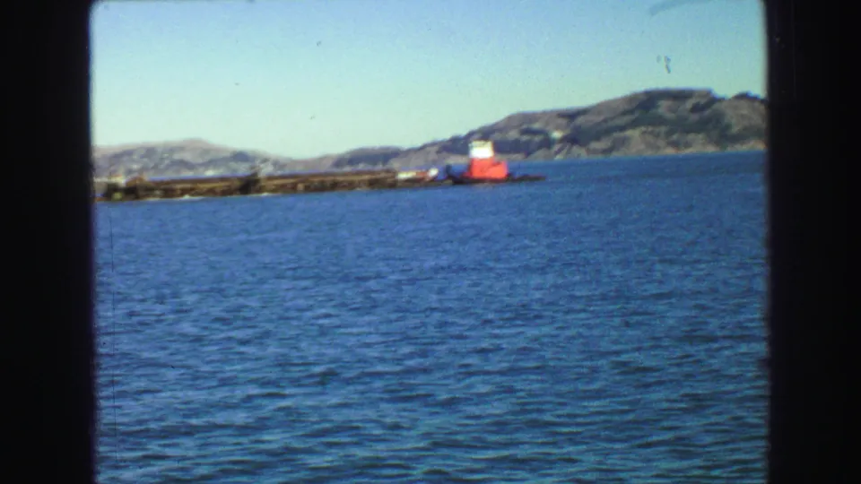 1968 Vancouver-area harbor view from a ferry, showing blue water, a low shoreline, hills and a red-and-white vessel or structure.
