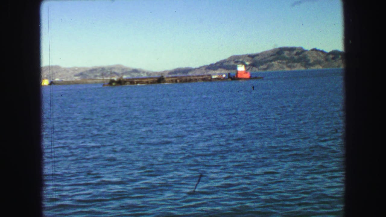 Blue water with a long harbor structure, red-and-white marker, hills, and clear sky.