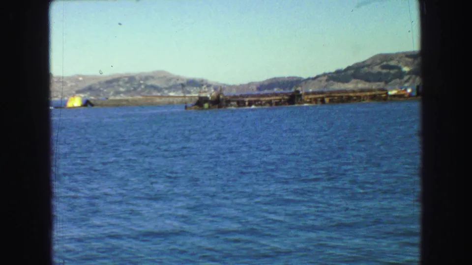 1968 Vancouver-area 8mm frame showing a yellow marker near a pier-like structure, with blue water and hills beyond.