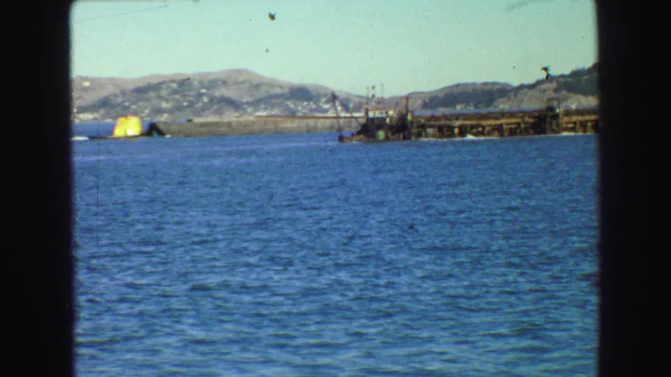 1968 Vancouver-area frame showing blue water, a yellow marker, a working pier or dock, distant shoreline, and hills.