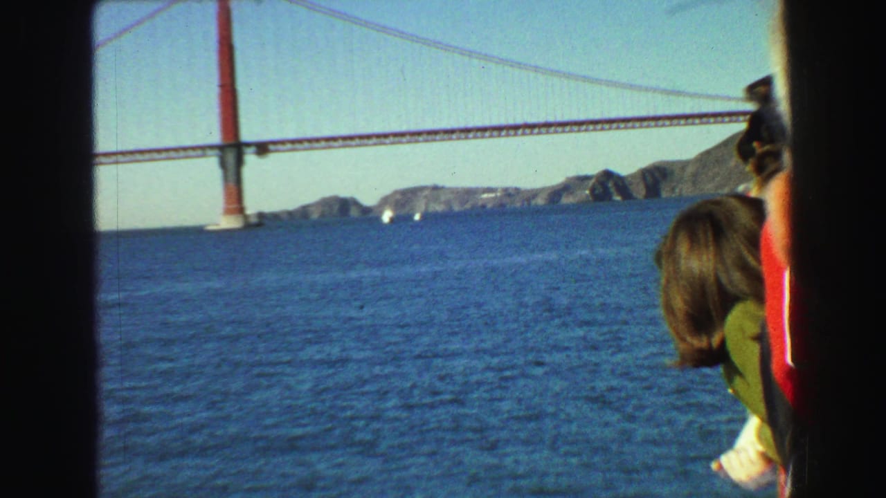 1968 8mm frame of passengers viewed from inside a boat as a red suspension bridge spans blue bay water in the distance.