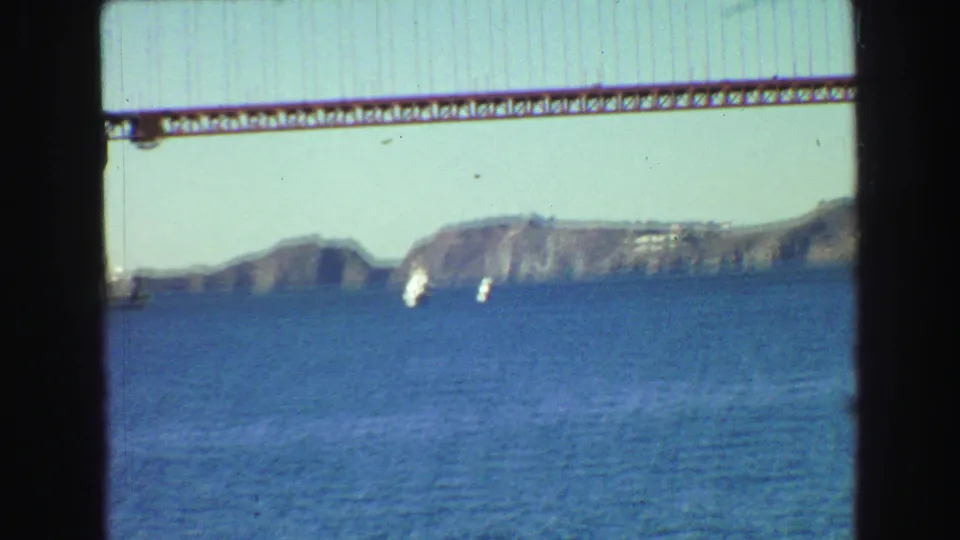 1968 8mm frame showing the bridge span above blue bay water, with rocky shoreline and small white boats in the distance.