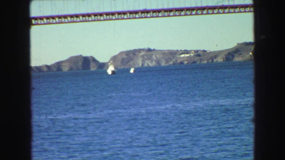 1968 8mm boat-view frame with blue bay water, distant rocky hills, small white boats, and the bridge span overhead.