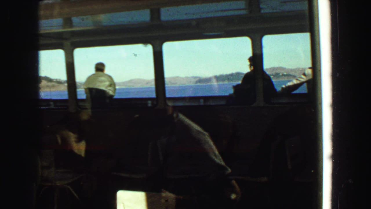 1968 Vancouver-area 8mm home-movie frame showing silhouetted passengers inside a boat bridge or cabin, with blue water and low coastline visible through broad windows.