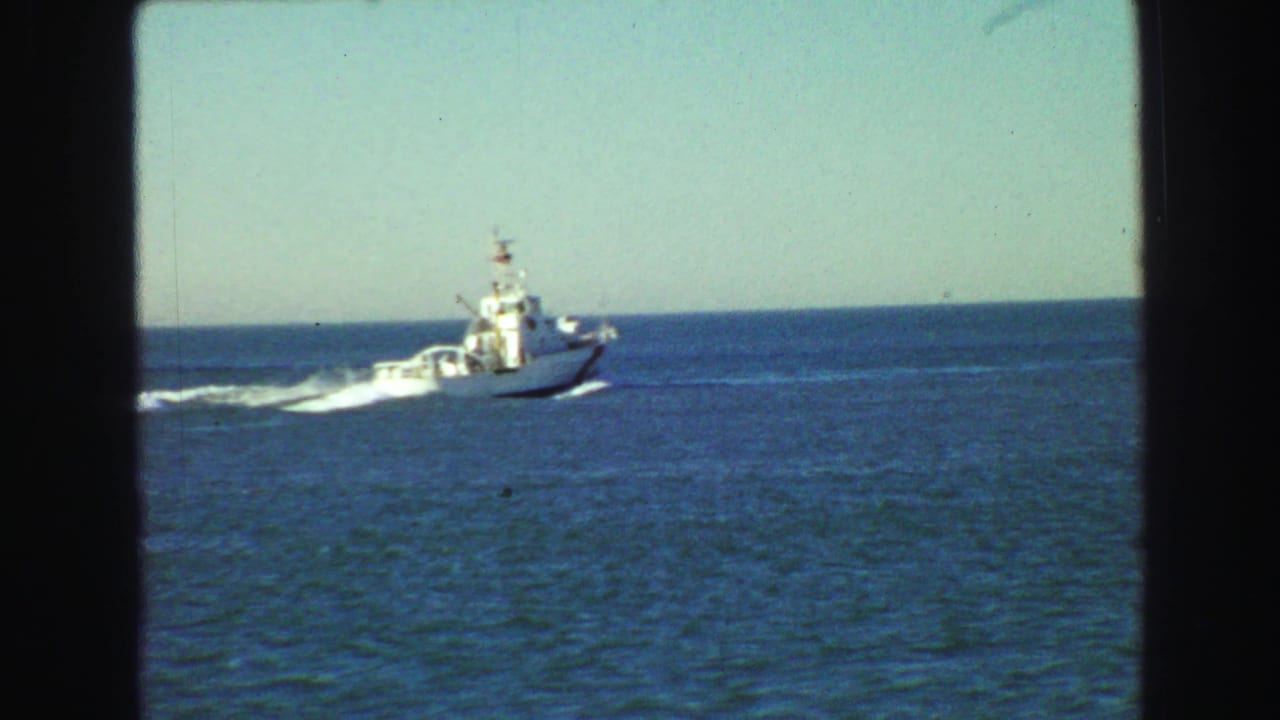 1968 Vancouver home-movie frame showing a white boat moving across calm blue water with a white wake behind it.