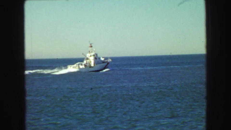 1968 Vancouver home-movie frame showing a white boat moving across calm blue water with a white wake behind it.